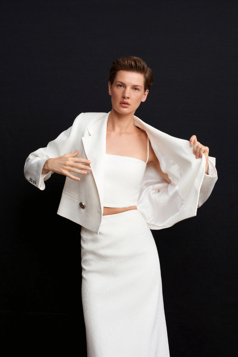 White Ancient Crop Blazer by Acaria Studio, stylish jacket with a modern design, displayed on a hanger against a plain background.