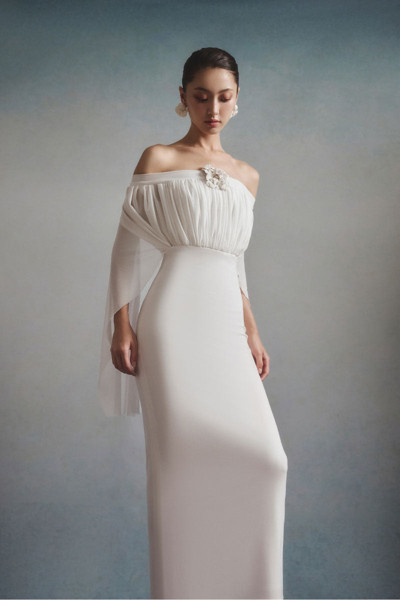 White Plan Elara off-shoulder maxi dress, flowing fabric with ruffled neckline, worn by a model in a scenic outdoor setting.