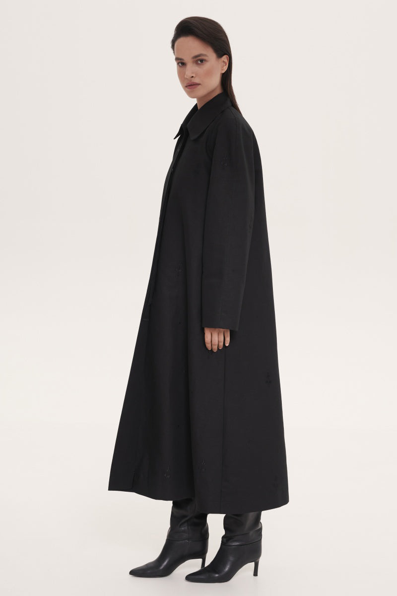 Black trench coat by Delegan hanging on a wooden hanger, showcasing its classic design and tailored fit.