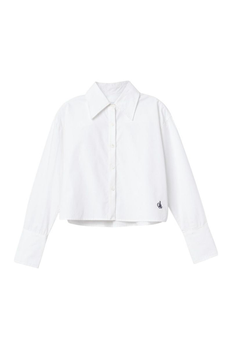 Dāl the Label cropped cotton button-down shirt with long sleeves and a classic collar, shown in a natural fabric texture.