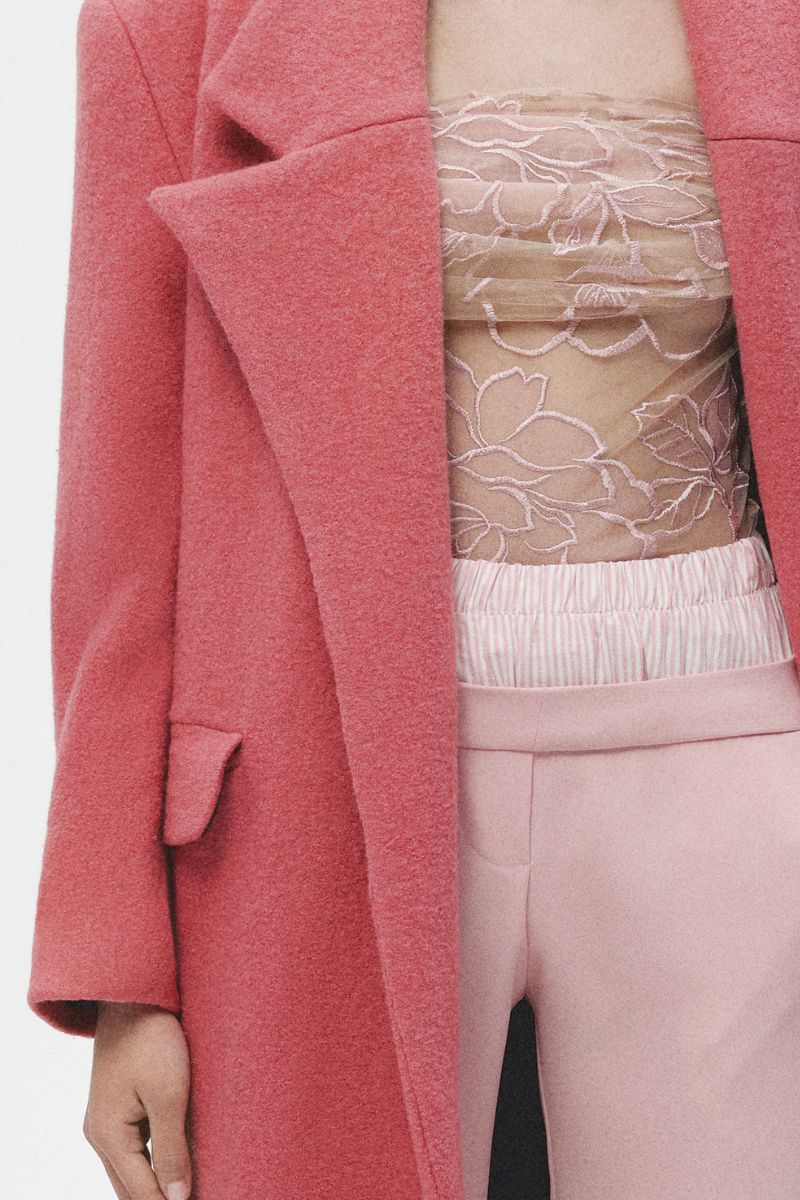 Pink wool maxi coat by JC Pajares hanging on a wooden hanger, showcasing its length and soft fabric texture.