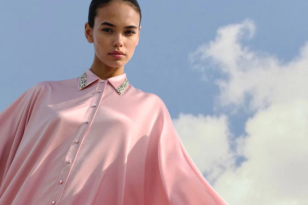 A model wearing a light pink satin cape-style button-up shirt with jeweled embellished collar, set against a blue sky with clouds.