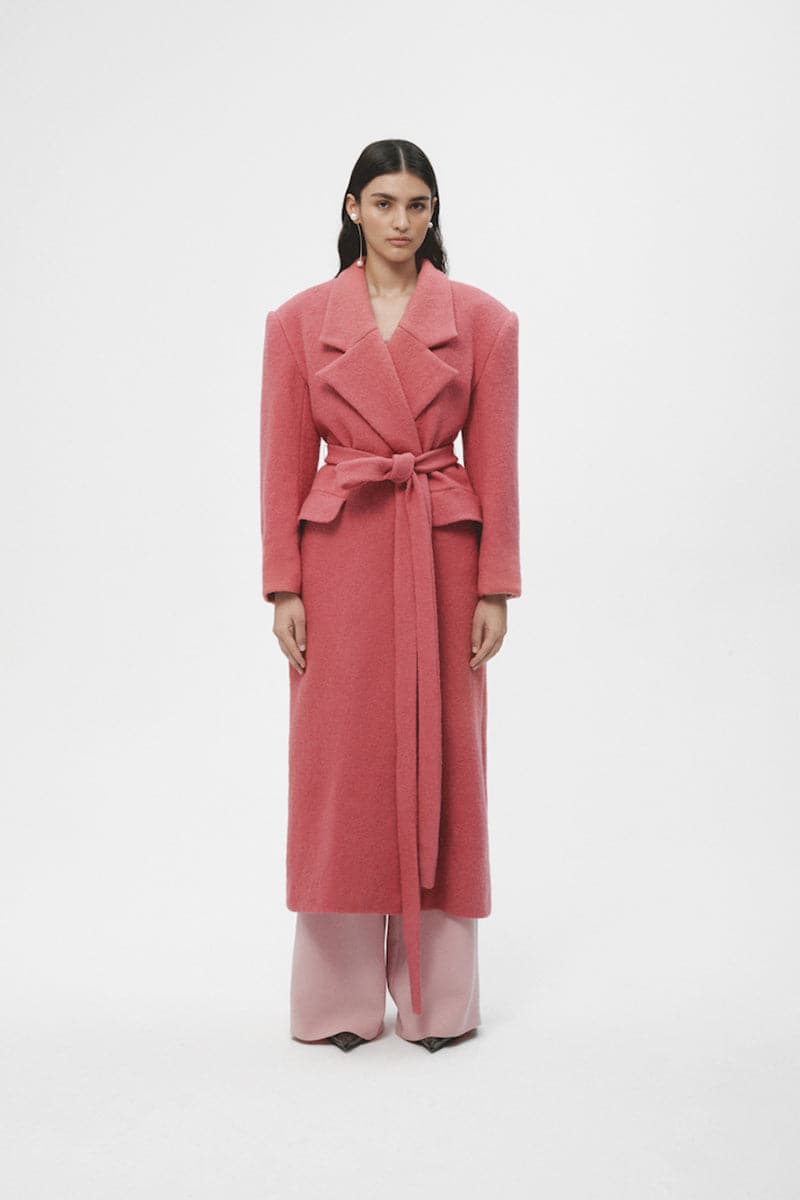 Pink wool maxi coat by JC Pajares hanging on a clothing rack, showcasing its long, cozy design and elegant texture.