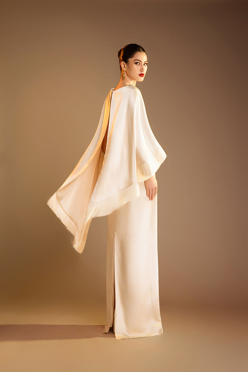 Cream column gown with fringed trim cape by Valenciani, featuring elegant flowing fabric and stylish layered design.