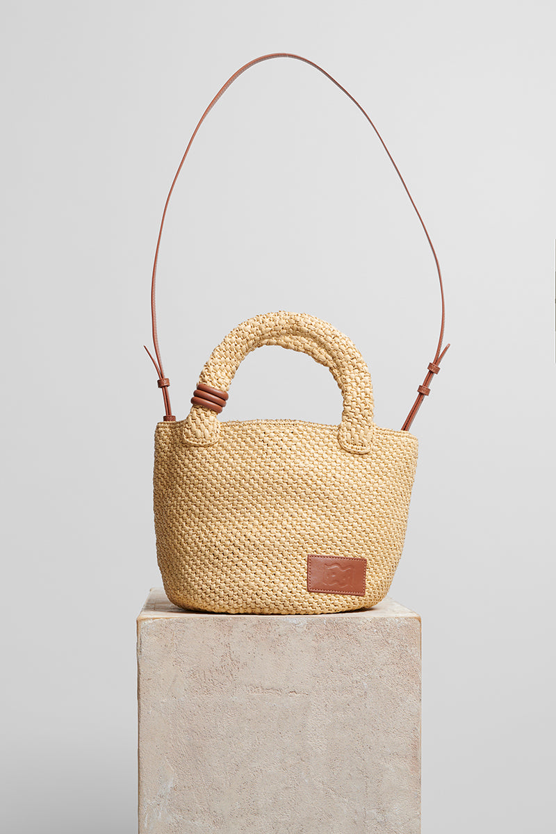 Beige raffia basket bag with top handle from Folklore The Label, showcasing a stylish and natural woven design.