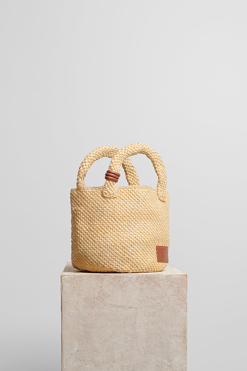 Beige raffia basket bag with top handle from Folklore The Label, featuring a woven design and natural texture.
