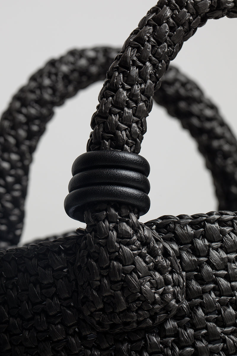 Black raffia basket bag with top handle from Folklore The Label, featuring a woven design and stylish, casual look.