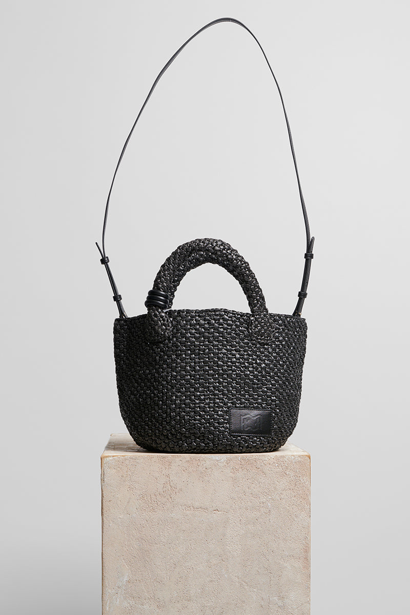 Black raffia basket bag with top handle from Folklore The Label, showcasing its woven texture and stylish design.