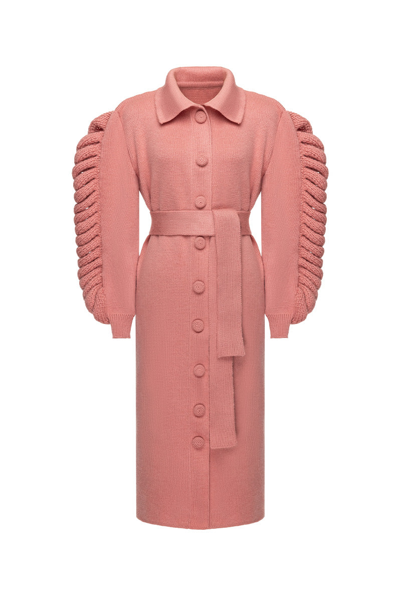 Liya dramatic pink knit belted coat hanging on a wooden background, featuring a textured knit fabric and a waist belt for style.