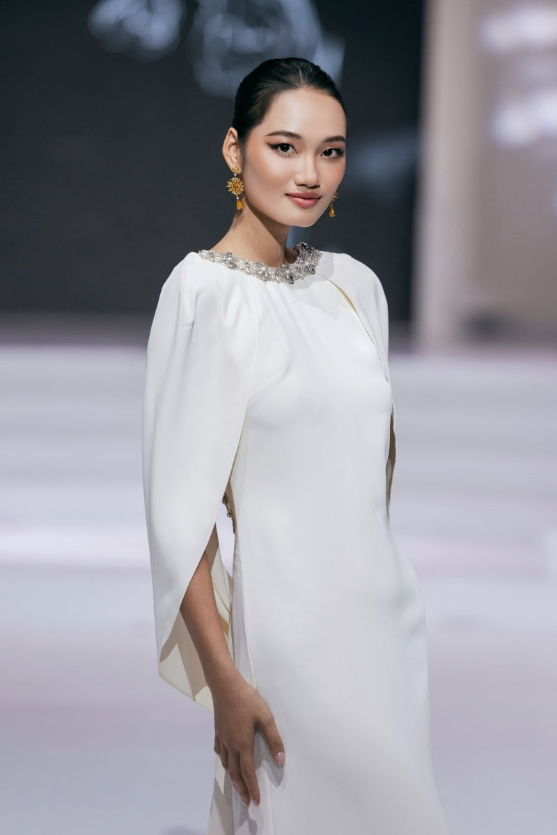 White Valenciani gown with a cowl back and cape sleeves, elegant evening dress on runway.