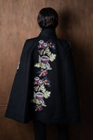 Nadya Shah Gaia Black Wisteria Cape jacket with flowing fabric and floral detailing, displayed on a hanger against a neutral background.