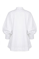 White FIORA top by ANA + MARIA displayed on a mannequin with a yellow magnolia blouse in the background.