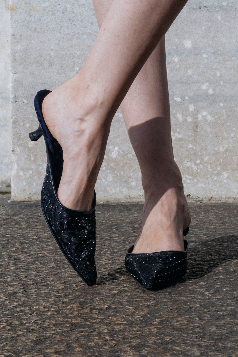 A pair of navy metal stud heels by AERA with pointed toes and ankle straps, displayed on a white background.