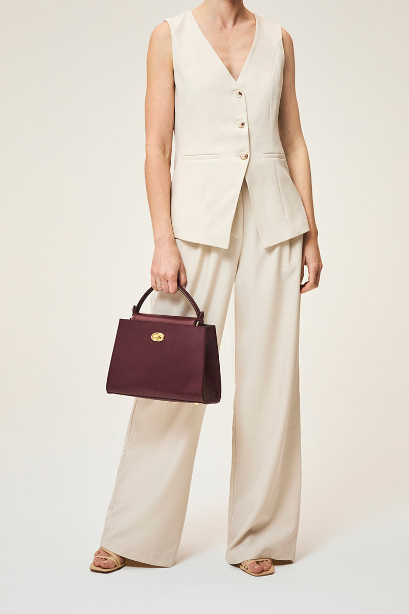 TUSTING Clara Grande Oxblood top handle bag with structured design and gold-tone hardware on smooth leather surface.