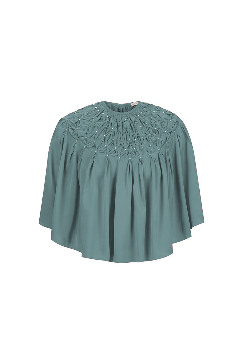 Hand-smocked TIYA cape in Saxon color by MASTANI, featuring intricate detailing and a stylish top design.