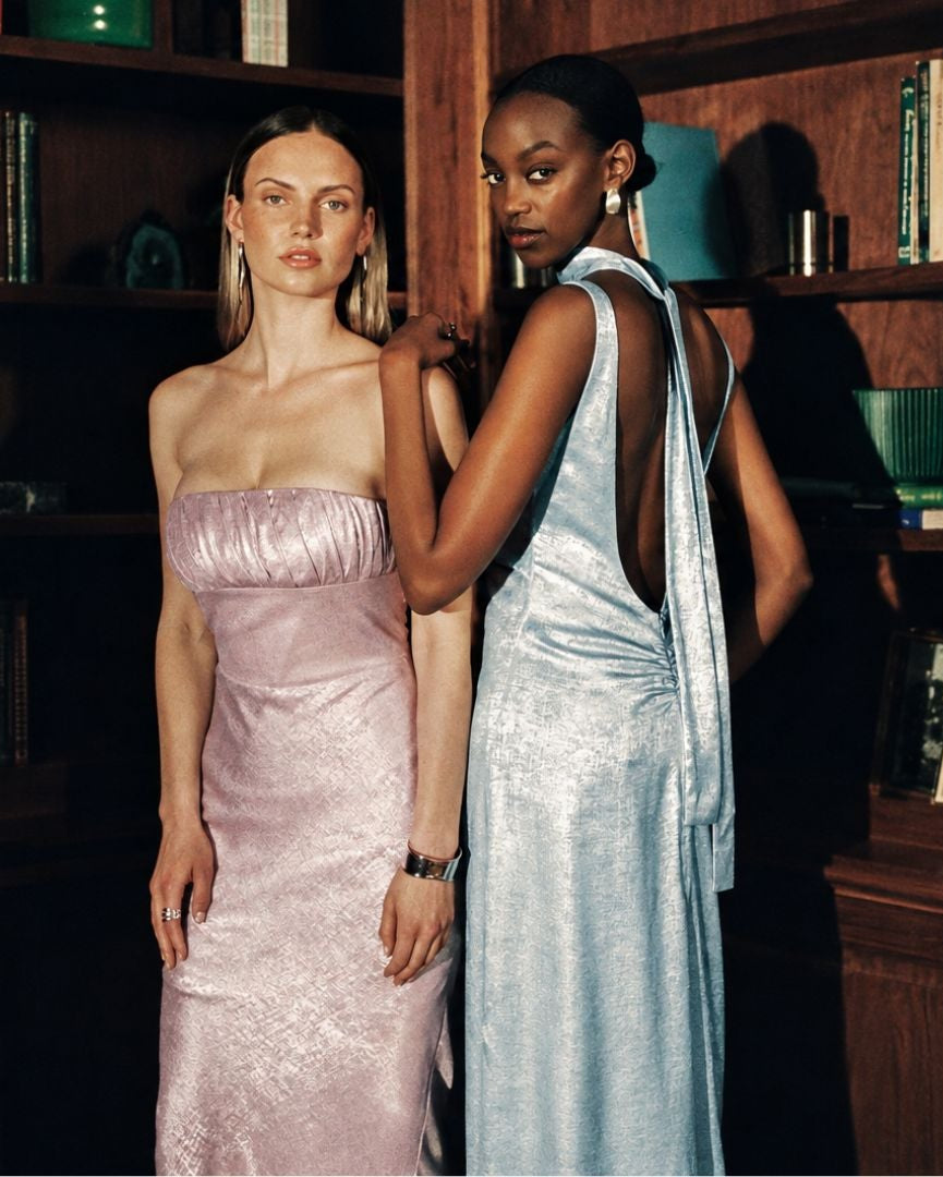 Two models wearing elegant evening gowns standing in front of dark wood bookshelves. The model on the left wears a strapless light pink satin gown, and the model on the right wears a light blue satin backless gown.