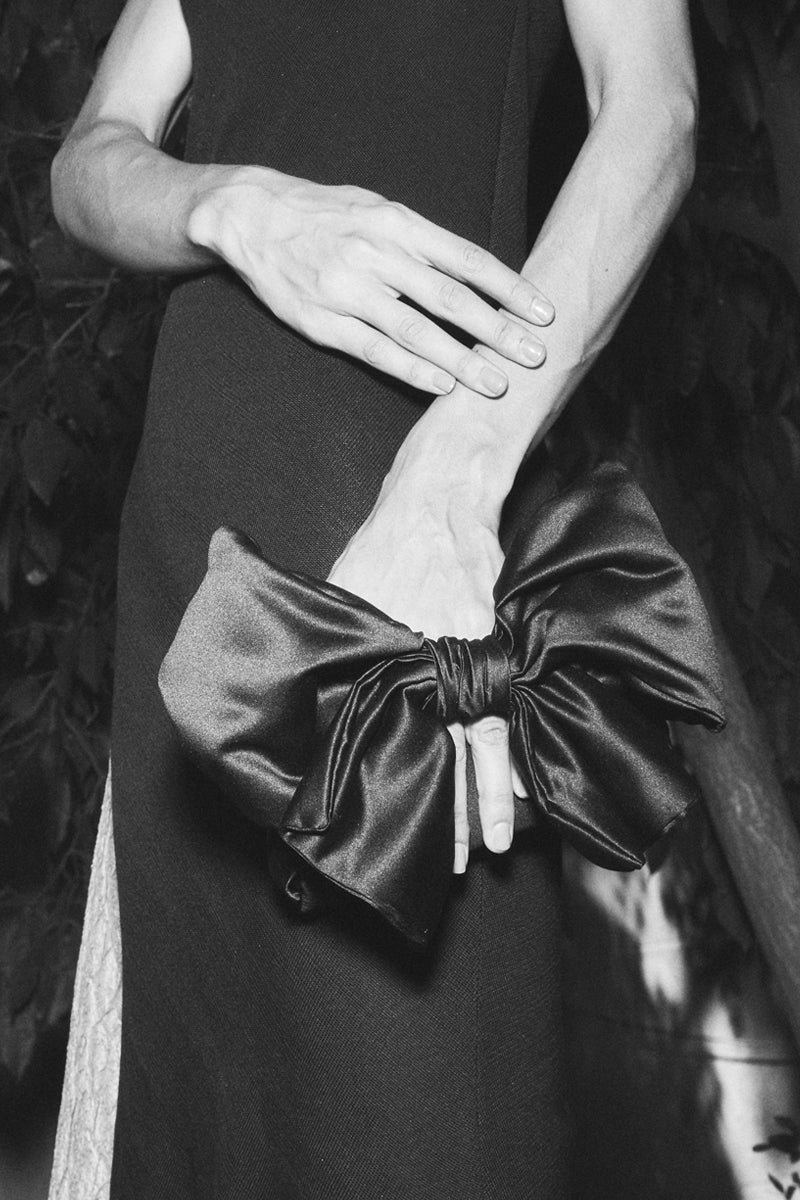 Black Sarah's Bag Bella handbag with a large bow, shoulder bag style, held by a model showcasing its elegant design.
