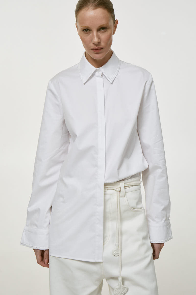 White embroidered shirt from DELEGAN featuring detailed embroidery, long sleeves, and a classic fit, displayed on a hanger.