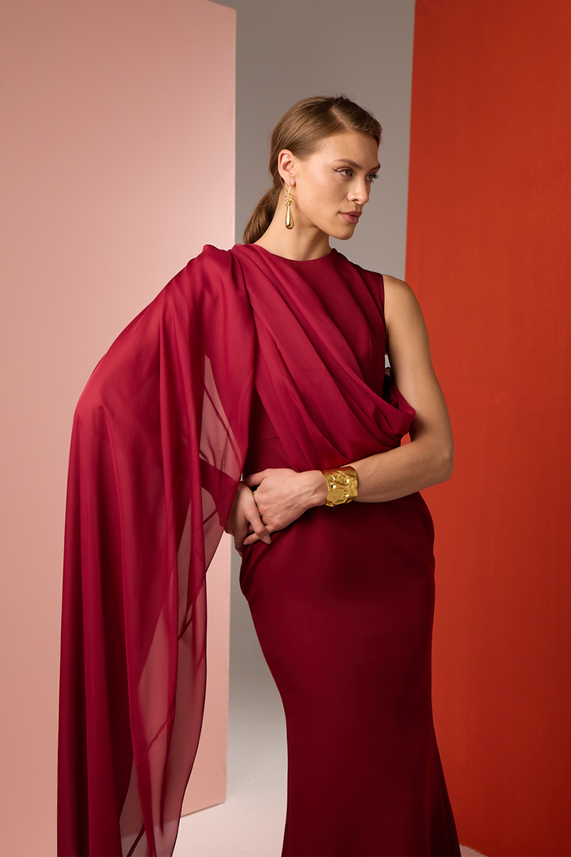 Elegant burgundy one-shoulder cape gown by Joanna Andraos, featuring flowing fabric and sophisticated design.