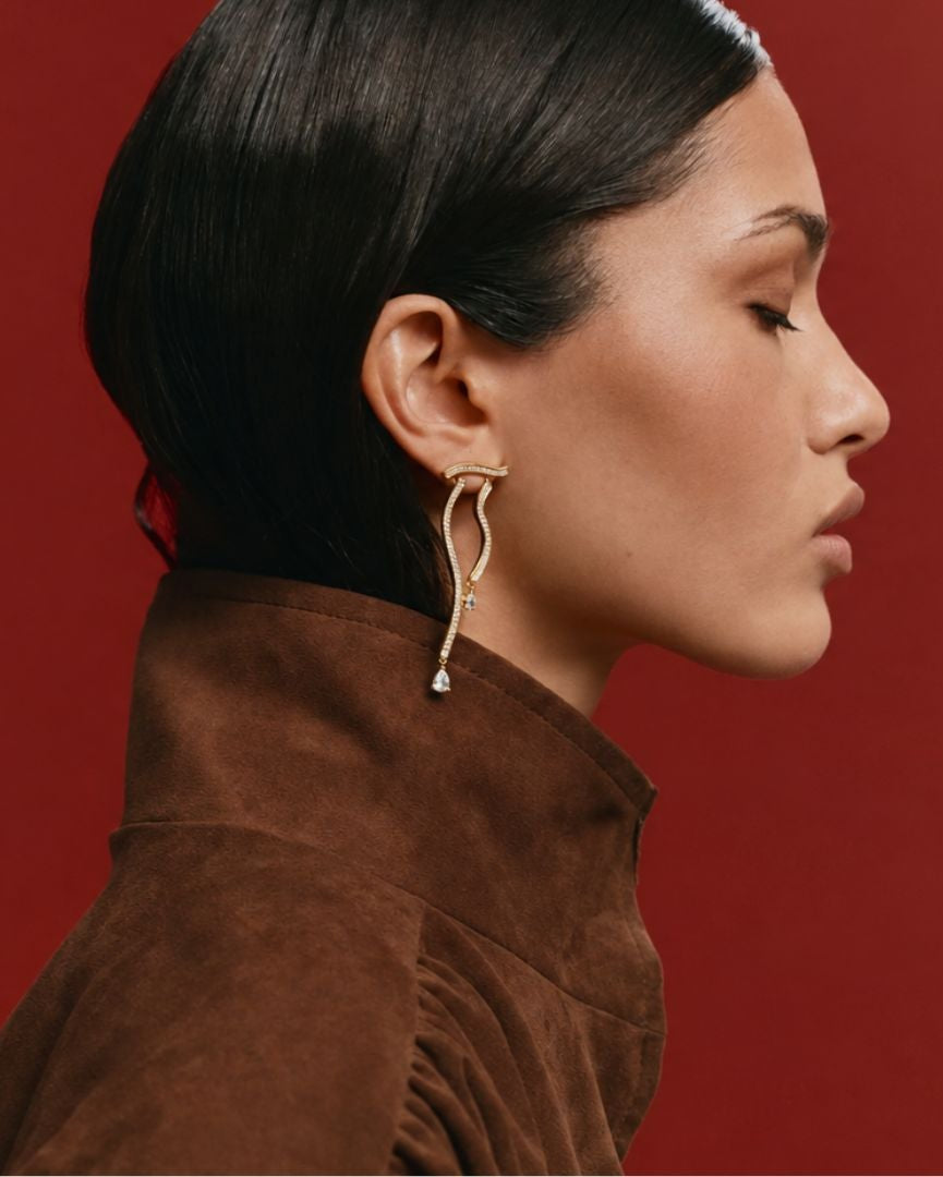 A woman in profile wearing a brown suede jacket and a gold dangle earring with a small clear stone, set against a solid dark red background.