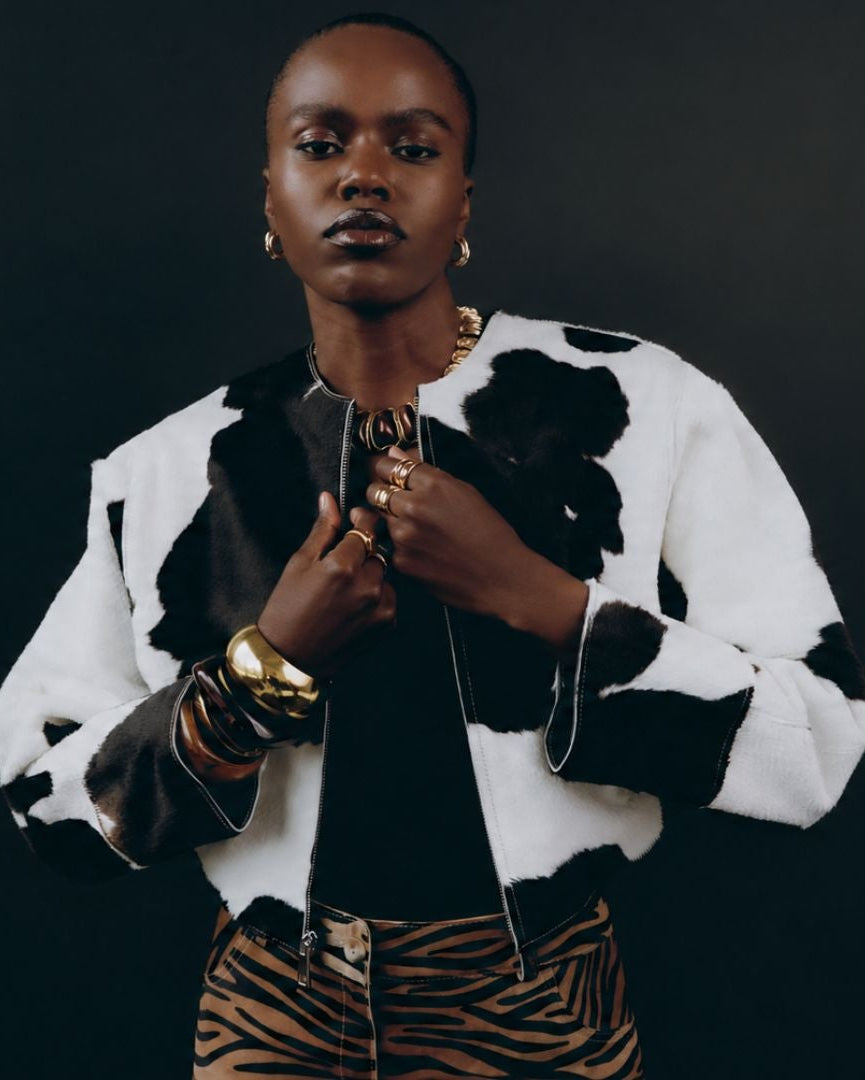 A model wearing a black and white cow-print jacket over a black top and tiger-print pants, accessorized with gold jewelry, set against a dark studio background.